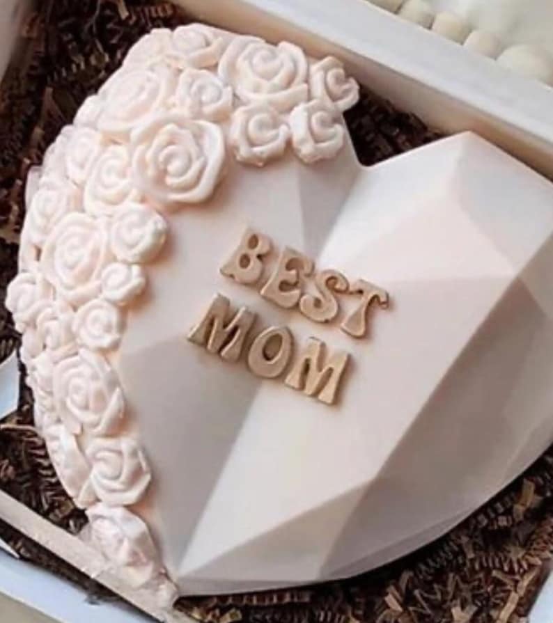 May include: A white heart-shaped cake decorated with white roses and the words "BEST MOM" in gold letters.