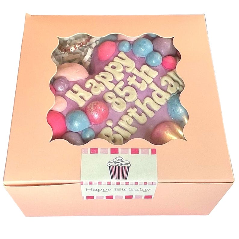 May include: A pink heart-shaped chocolate gift box with a clear window showing a purple chocolate heart with the text "Happy 85th Birthday" in white letters and colorful sprinkles. The box has a small label on the front that says "Happy Birthday" with a cupcake graphic.