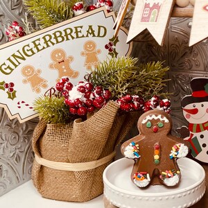 May include: A gingerbread themed Christmas decoration with a burlap wrapped pot filled with greenery and red berries. A chocolate gingerbread man with sprinkles is on a white pedestal.