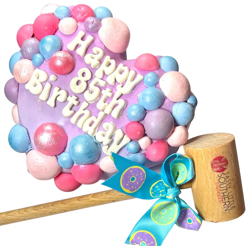 May include: Purple heart-shaped cake decorated with pink, blue, and white frosting and sprinkles. The cake says "Happy 85th Birthday". The cake is on a wooden mallet with a blue ribbon with donut designs.