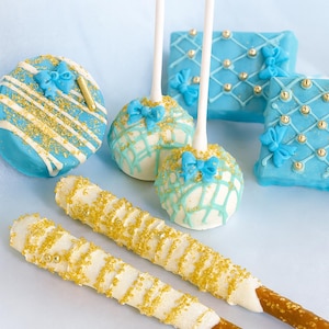 Pu&ograve; includere: Un assortimento di dolcetti decorati, tra cui cake pops blu e bianchi con fiocchi, dolci rettangolari blu con dettagli dorati e bastoncini di pretzel ricoperti di cioccolato bianco e granella dorata. Disposti su una superficie azzurra.