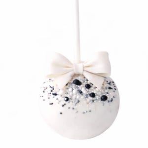 Puede incluir: Una paleta de pastel blanca con un lazo blanco en la parte superior, decorada con chispas blancas, plateadas y negras. La paleta de pastel est&aacute; en un palito blanco, ideal para una fiesta.