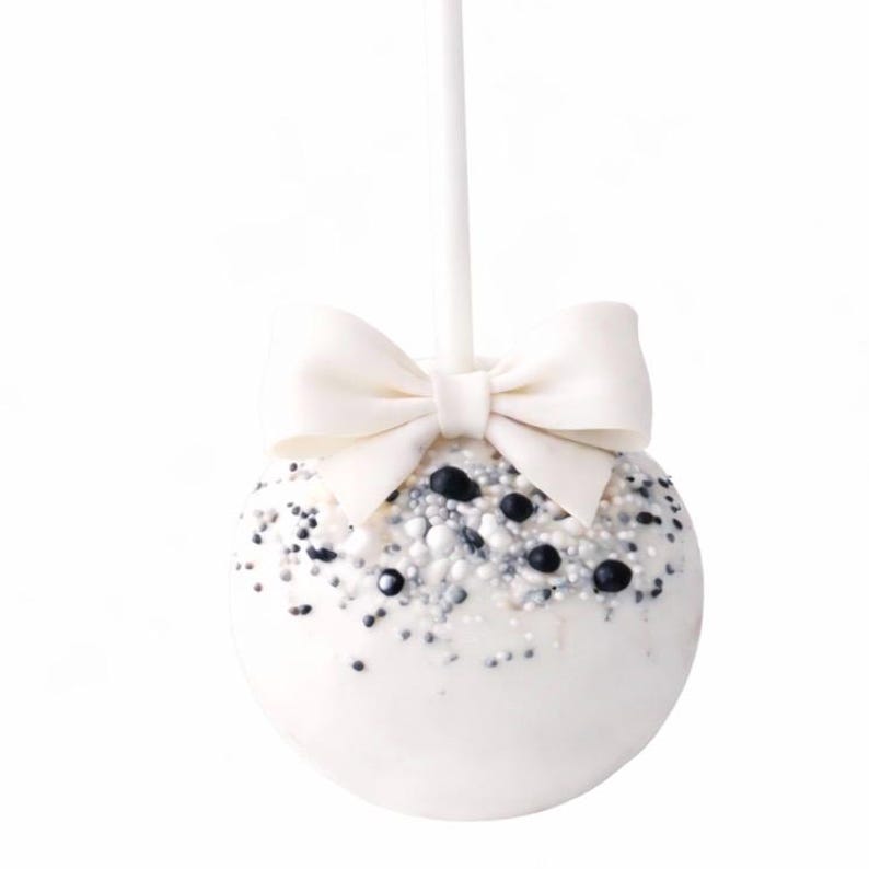 Puede incluir: Una paleta de pastel blanca con un lazo blanco en la parte superior, decorada con chispas blancas, plateadas y negras. La paleta de pastel est&aacute; en un palito blanco, ideal para una fiesta.