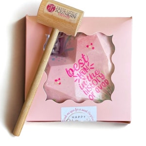 May include: A pink heart-shaped chocolate bomb with the text "best mom in the history of ever" and a small wooden hammer to break it open. The box is pink with a white border and the text "Break me open for a sweet surprise" and "Happy"