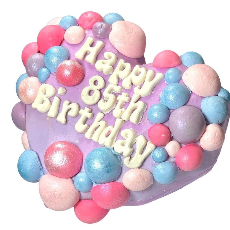 May include: Purple heart-shaped cake decorated with pastel pink, blue, and purple frosting balls. The cake is decorated with white frosting lettering that says "Happy 85th Birthday".