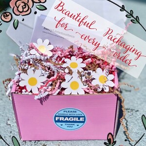 K&ouml;nnte beinhalten: Rosa Geschenkbox gef&uuml;llt mit braunem und rosa Knisterpapier und wei&szlig;en Papierblumen in Form von G&auml;nsebl&uuml;mchen. Die Box hat einen blau-wei&szlig;en Aufkleber mit der Aufschrift "Bitte vorsichtig behandeln Zerbrechlich Danke".