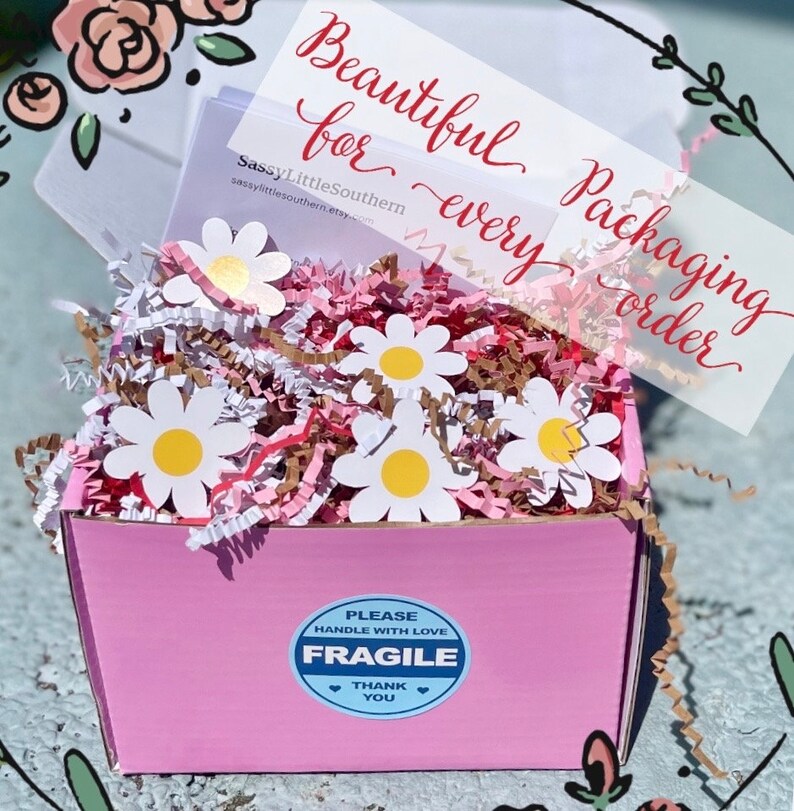K&ouml;nnte beinhalten: Rosa Geschenkbox gef&uuml;llt mit braunem und rosa Knisterpapier und wei&szlig;en Papierblumen in Form von G&auml;nsebl&uuml;mchen. Die Box hat einen blau-wei&szlig;en Aufkleber mit der Aufschrift "Bitte vorsichtig behandeln Zerbrechlich Danke".