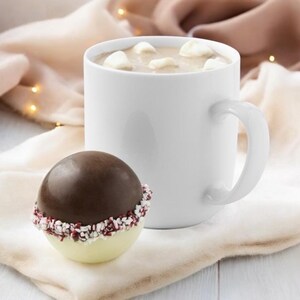 Puede incluir: Una taza blanca llena de chocolate caliente cubierto con malvaviscos junto a una bomba de chocolate caliente de chocolate y menta blanca.