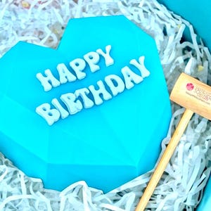May include: A blue heart-shaped cake with white letters spelling "Happy Birthday" on top. The cake is sitting in a white shredded paper gift box.