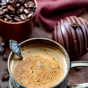 K&ouml;nnte beinhalten: Nahaufnahme einer Tasse Kaffee mit einem silbernen L&ouml;ffel darin. Der Kaffee ist schaumig und hat eine dunkelbraune Farbe. Die Tasse ist aus Metall und hat einen abgenutzten Look. Im Hintergrund befindet sich eine Schokoladentr&uuml;ffel.