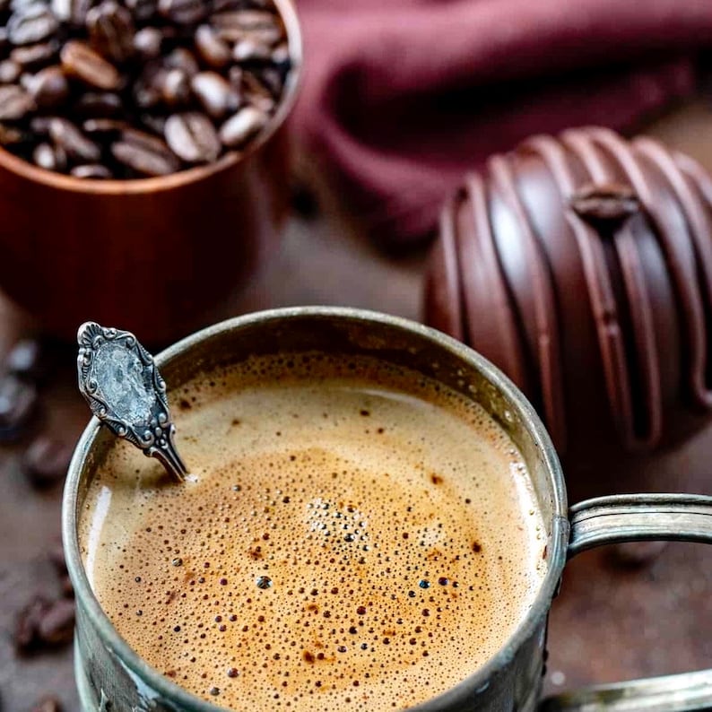 K&ouml;nnte beinhalten: Nahaufnahme einer Tasse Kaffee mit einem silbernen L&ouml;ffel darin. Der Kaffee ist schaumig und hat eine dunkelbraune Farbe. Die Tasse ist aus Metall und hat einen abgenutzten Look. Im Hintergrund befindet sich eine Schokoladentr&uuml;ffel.