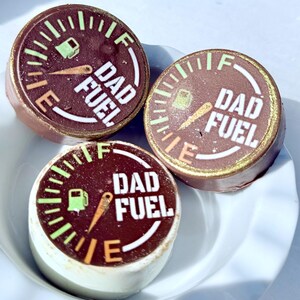 May include: Three round chocolate treats with a gauge design and the words "DAD FUEL" in white. The gauge features a fuel pump icon and an orange needle. The treats are arranged in a white bowl.