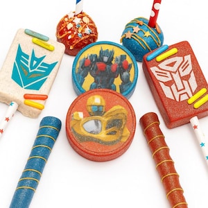 May include: An assortment of colorful cake pops and rectangular treats on sticks. The treats feature images of Transformers characters and logos in red, blue, yellow, and white. The sticks are decorated with stars and stripes.
