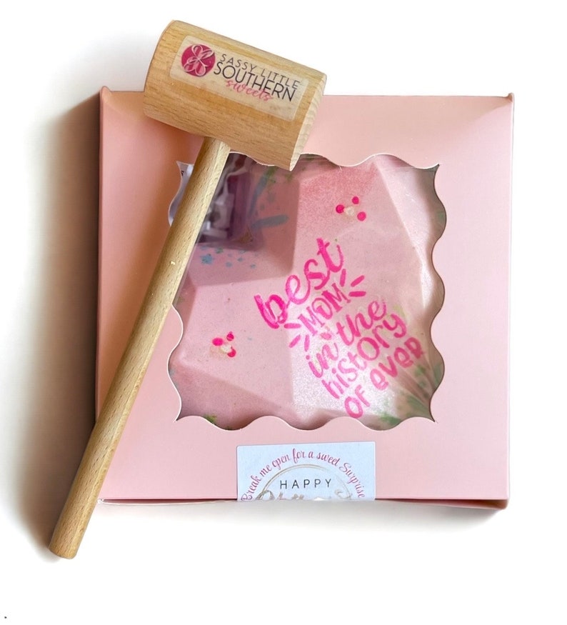 May include: A pink heart-shaped chocolate bomb with the text "best mom in the history of ever" and a small wooden hammer to break it open. The box is pink with a white border and the text "Break me open for a sweet surprise" and "Happy"