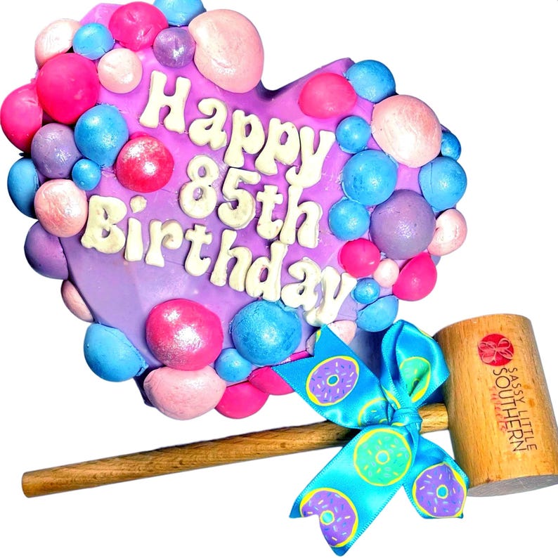 May include: A purple heart-shaped cake decorated with colorful sprinkles and the words "Happy 85th Birthday". A wooden mallet with a blue ribbon is next to the cake.