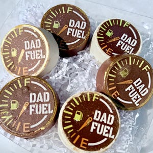 May include: Six round chocolate treats with a gauge design and the words "DAD FUEL" in white. The treats have a brown and white color scheme with gold accents. The treats are arranged in a clear box with white paper filler.