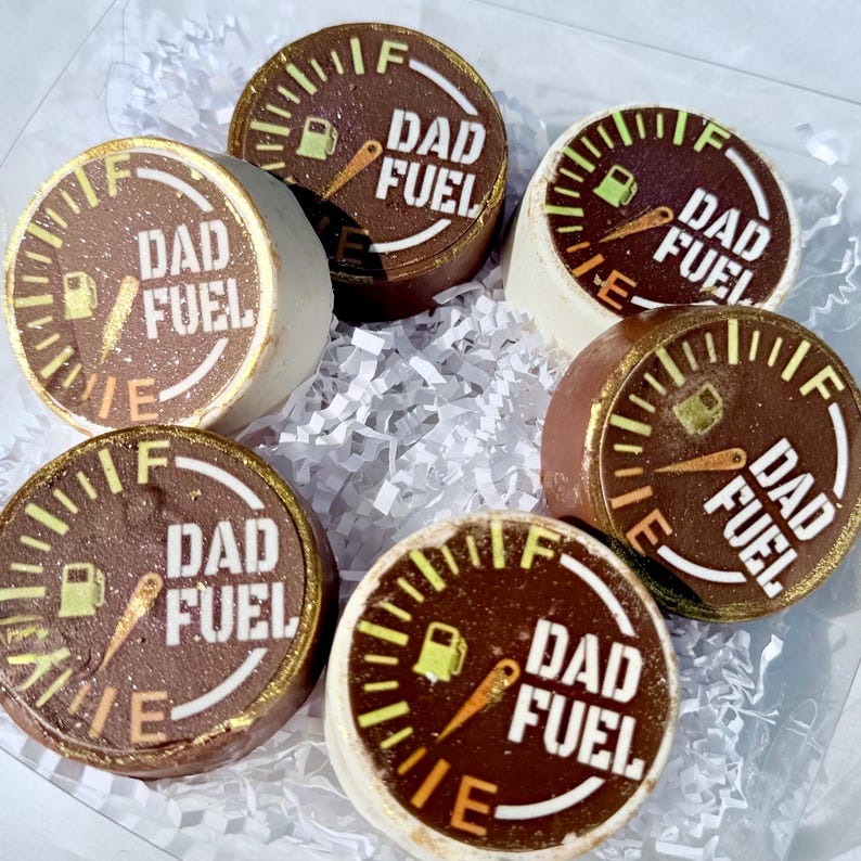 May include: Six round chocolate treats with a gauge design and the words "DAD FUEL" in white. The treats have a brown and white color scheme with gold accents. The treats are arranged in a clear box with white paper filler.