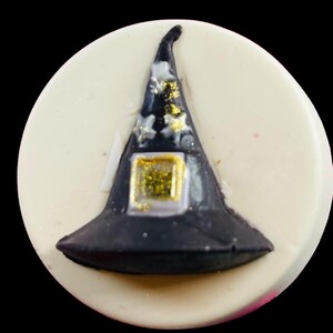 Puede incluir: Un dulce tem&aacute;tico de Halloween con un sombrero de bruja negro y detalles dorados, sobre un fondo blanco circular. El sombrero tiene una hebilla cuadrada dorada. Probablemente sea una galleta o un caramelo.