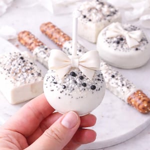 Puede incluir: Una paleta de pastel blanca con un lazo crema, decorada con chispas plateadas y negras. Otros dulces incluyen pretzels cubiertos de chocolate blanco y pasteles cuadrados, todos decorados de manera similar. Los dulces est&aacute;n sobre una superficie blanca.