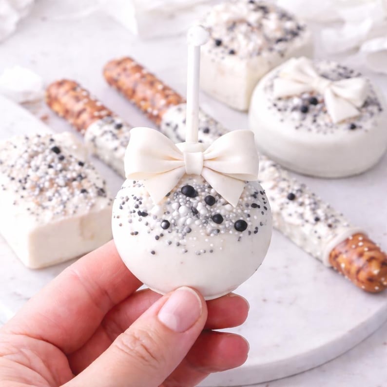Puede incluir: Una paleta de pastel blanca con un lazo crema, decorada con chispas plateadas y negras. Otros dulces incluyen pretzels cubiertos de chocolate blanco y pasteles cuadrados, todos decorados de manera similar. Los dulces est&aacute;n sobre una superficie blanca.