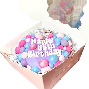 May include: A purple heart-shaped cake decorated with pink, blue, and white sprinkles and the words "Happy 85th Birthday". The cake is in a pink box with white tissue paper.