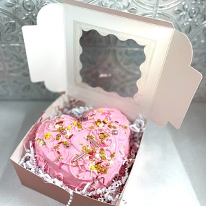 May include: A pink heart-shaped cake decorated with sprinkles and gold accents is displayed in a pink box with a clear window.