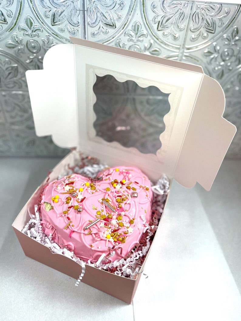 May include: A pink heart-shaped cake decorated with sprinkles and gold accents is displayed in a pink box with a clear window.
