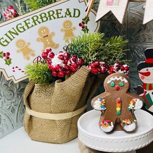 May include: A gingerbread man cookie decorated with colorful sprinkles and white frosting. The cookie is sitting on a white cake stand with a burlap base. The background includes a sign that says "Gingerbread" with a green background and red berries.