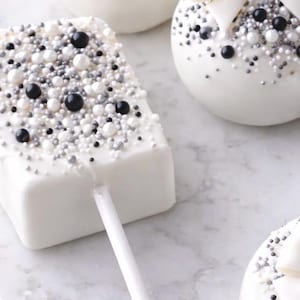 Puede incluir: Cake pops rectangulares blancos sobre palito, cubiertos de chocolate blanco y decorados con chispas negras, plateadas y blancas. Otros cake pops blancos con decoraciones similares est&aacute;n en el fondo. Los dulces est&aacute;n sobre una superficie de m&aacute;rmol.