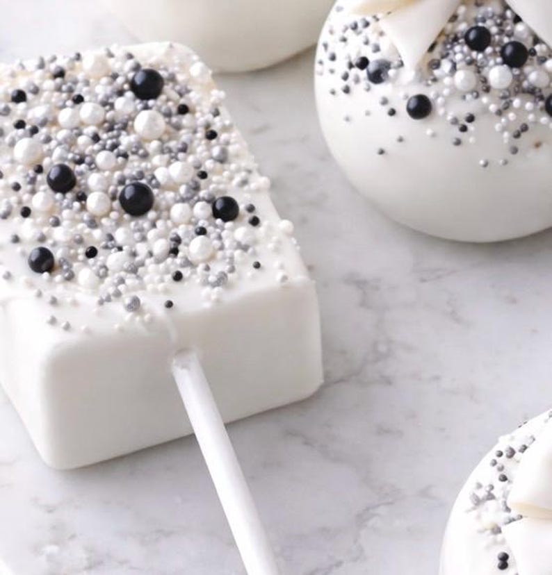 Puede incluir: Cake pops rectangulares blancos sobre palito, cubiertos de chocolate blanco y decorados con chispas negras, plateadas y blancas. Otros cake pops blancos con decoraciones similares est&aacute;n en el fondo. Los dulces est&aacute;n sobre una superficie de m&aacute;rmol.