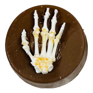 Puede incluir: Un dulce redondo de chocolate negro, coronado con una mano de esqueleto blanca. La mano tiene detalles dorados y est&aacute; colocada como si emergiera del chocolate. Un dulce tem&aacute;tico de Halloween.