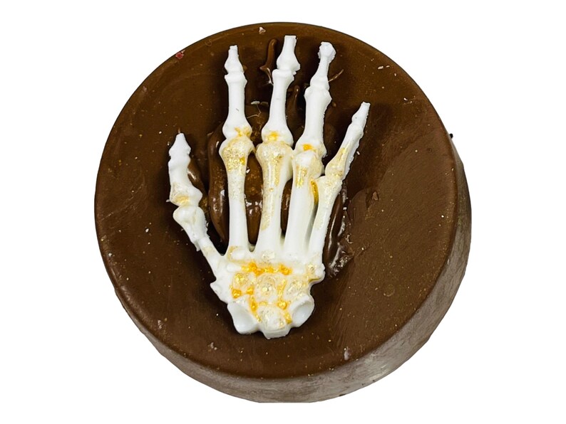 Puede incluir: Un dulce redondo de chocolate negro, coronado con una mano de esqueleto blanca. La mano tiene detalles dorados y est&aacute; colocada como si emergiera del chocolate. Un dulce tem&aacute;tico de Halloween.