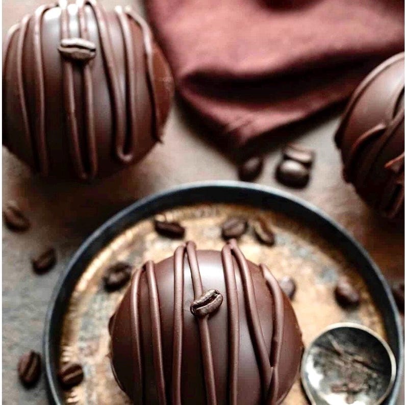 K&ouml;nnte beinhalten: Drei Schokoladenbomben mit Kaffeebohnen darauf. Die Schokoladenbomben sind mit Schokoladenstreifen verziert. Die Bomben befinden sich auf einer Holzoberfl&auml;che mit einem braunen Tuch im Hintergrund.
