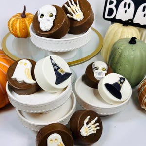 Puede incluir: Una colecci&oacute;n de golosinas de chocolate con tem&aacute;tica de Halloween. Las golosinas est&aacute;n decoradas con fantasmas blancos, sombreros de bruja, calaveras y manos de esqueleto. Tambi&eacute;n se ven calabazas y un letrero de "Boo".