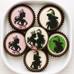 May include: A collection of decorated cupcakes in a white tray. The cupcakes feature black silhouettes of a princess and a witch on pink, white, and green backgrounds. Each cupcake is in a brown paper liner.