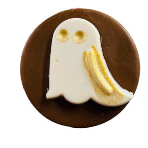 Puede incluir: Un caramelo de chocolate redondo con un dise&ntilde;o de fantasma blanco. El fantasma tiene ojos dorados y detalles dorados en los lados. Un dulce tem&aacute;tico de Halloween.