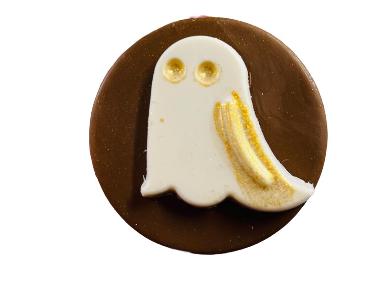 Puede incluir: Un caramelo de chocolate redondo con un dise&ntilde;o de fantasma blanco. El fantasma tiene ojos dorados y detalles dorados en los lados. Un dulce tem&aacute;tico de Halloween.