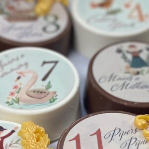 Puede incluir: Un conjunto de favores redondos de chocolate con diferentes dise&ntilde;os y n&uacute;meros. Cada favor tiene una base de chocolate blanco con un dise&ntilde;o colorido y un acento dorado. Los dise&ntilde;os incluyen un cisne, un patr&oacute;n floral y una mujer con un vestido. Los n&uacute;meros en los favores van del 1 al 8.