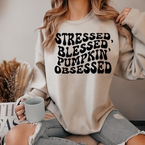 May include: A beige sweatshirt with the text "Stressed, Blessed, Pumpkin' Obsessed" in black, retro-style lettering.