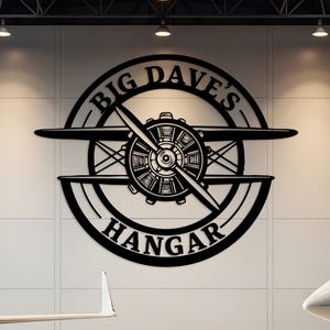 May include: A black metal sign with the text "Big Dave's Hangar" and a propeller design is mounted on a white wall. Two white private jets are parked in a hangar with a reflective floor.