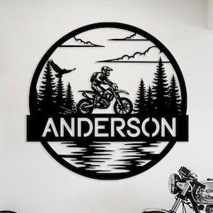 Puede incluir: Arte de pared de metal negro con un piloto de motocross en una motocicleta, dentro de un diseño circular con árboles, agua y nubes. El nombre "ANDERSON" se muestra de forma destacada. Una motocicleta y una rueda son visibles en el fondo.