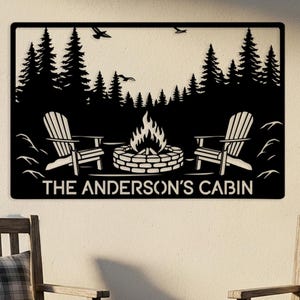 May include: Black metal wall art featuring a cabin scene with a campfire, Adirondack chairs, and trees. The text "THE ANDERSON'S CABIN" is displayed below the scene. Two wooden chairs with plaid cushions and a wooden stump with pine cones and a mug are in the foreground.