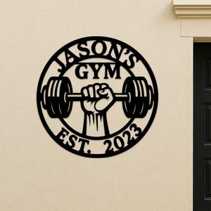 May include: Black metal wall art with the text "JASON'S GYM EST. 2023" in a circular design. A hand is lifting a barbell in the center of the circle. The art is mounted on a beige wall next to a black door.