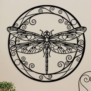 May include: Black metal dragonfly wall art. The dragonfly is centered within a circular frame with decorative swirls. The wings and body of the dragonfly are detailed with intricate patterns. Suitable for indoor or outdoor use.