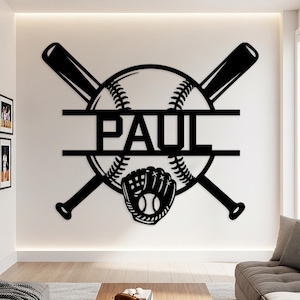 Op de afbeelding: Zwarte honkbalmuurkunst met een honkbal, gekruiste knuppels en een handschoen. De naam "PAUL" wordt in het midden weergegeven. De kunst is gemonteerd op een witte muur in een woonkamer.