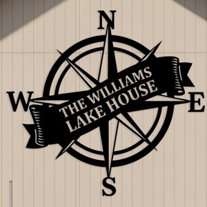 Pode incluir: Uma placa de rosa dos ventos de metal preto com o texto "THE WILLIAMS LAKE HOUSE" em uma faixa. A placa é montada em uma fachada de edifício bege claro. A bússola aponta para as direções cardeais: Norte, Sul, Leste e Oeste.