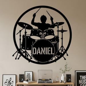 Op de afbeelding: Zwarte metalen wandkunst met een drummer die een drumstel bespeelt in een cirkel. De naam "DANIEL" staat op de basdrum. Een gitaar, ingelijste kunst en een platenspeler zijn eronder zichtbaar.