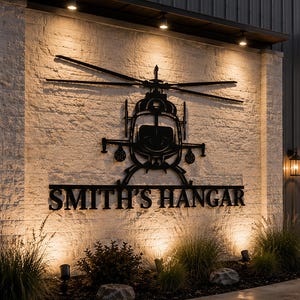 May include: A black metal helicopter silhouette is mounted on a textured, off-white brick wall. The text "SMITH'S HANGAR" is below the helicopter. The scene is illuminated by spotlights and a wall-mounted lantern.
