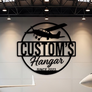 May include: Black metal sign with the text "CUSTOM'S Hangar SINCE 2023" and a silhouette of a small airplane. The sign is mounted on a white wall in a hangar with two airplanes.