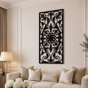 May include: A black, ornate wall panel with a symmetrical floral design is mounted on a cream-colored wall. The panel is rectangular and features intricate scrollwork and a central circular motif. The panel is a decorative element in a living room setting.